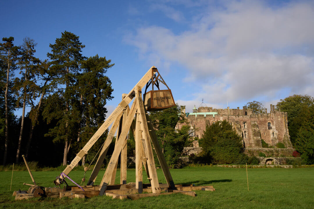 A stationary trebuchet in focus on the left, showcasing an overcast view of Berkeley Castle during the day in the background.
